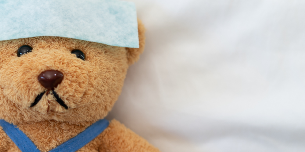 Sick brown teddy bear lying in bed with a towel over his head