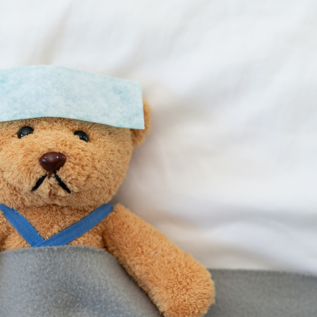 Sick brown teddy bear lying in bed with a towel over his head